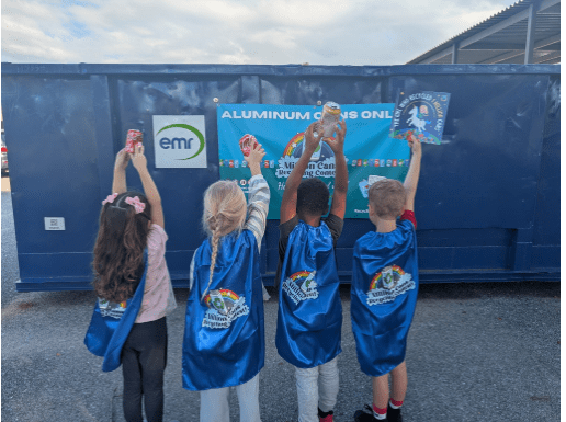 Recycling Society Million Cans Recycling Contest Four Elementary Students wearing superhero capes holding aluminum cans and the children's book, "The Girl Who Recycled a Million Cans" in front of an EMR metal recycling roll off container.